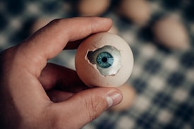 A hand is holding an eggshell that has an eye emerging from the cracked surface. The background is blurred, showcasing more egg-like shapes.