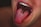 A close-up of a person's open mouth with several nails protruding from the tongue, creating a surreal and unsettling visual effect. The teeth are visible, and the background is dark with a subtle emphasis on the tongue and nails.