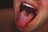 A close-up of a person's open mouth with several nails protruding from the tongue, creating a surreal and unsettling visual effect. The teeth are visible, and the background is dark with a subtle emphasis on the tongue and nails.