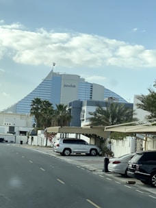 A large, modern building with a distinctive sail-like shape and reflective glass surfaces is seen in the background. The building is labeled 'Jumeirah' and is situated behind a row of palm trees. A street with parked cars and residential structures is visible in the foreground, with a partly cloudy sky above.