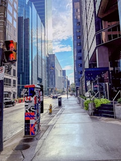 A city street lined with tall modern buildings, with a mix of glass and concrete facades. A traffic light displays a red hand, while the sky is partly cloudy with some blue patches. There is a decorated utility box near the sidewalk, beside a fire hydrant.