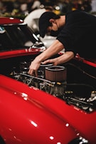 A person wearing a black cap and black shirt is working on the engine of a red car, adjusting parts within the engine compartment. The setting appears to be indoors, possibly a workshop or an event with other vehicles or people in the background.