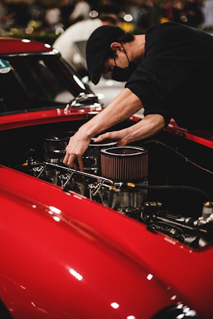 A person wearing a black cap and black shirt is working on the engine of a red car, adjusting parts within the engine compartment. The setting appears to be indoors, possibly a workshop or an event with other vehicles or people in the background.