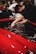 A person wearing a black cap and black shirt is working on the engine of a red car, adjusting parts within the engine compartment. The setting appears to be indoors, possibly a workshop or an event with other vehicles or people in the background.