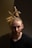 A person with light skin, a beard, and dreadlocked hair styled up in a ponytail is captured against a dark background. The person is wearing a black shirt and has a focused, contemplative expression.
