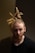 A person with light skin, a beard, and dreadlocked hair styled up in a ponytail is captured against a dark background. The person is wearing a black shirt and has a focused, contemplative expression.