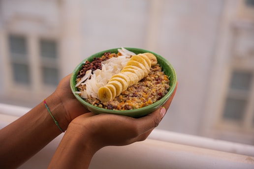 A person holds a green bowl filled with a mixture of grains and fresh toppings. The toppings include sliced banana, coconut flakes, and what appears to be cacao nibs or dried fruit. The bowl is supported by two hands, and the background is softly blurred, indicating an outdoor setting.