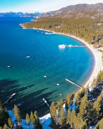 A serene beach with clear turquoise waters lined by a dense forest of evergreen trees. Piers extend into the water, and small boats are scattered across the surface. In the background, snow-capped mountains add to the picturesque landscape.