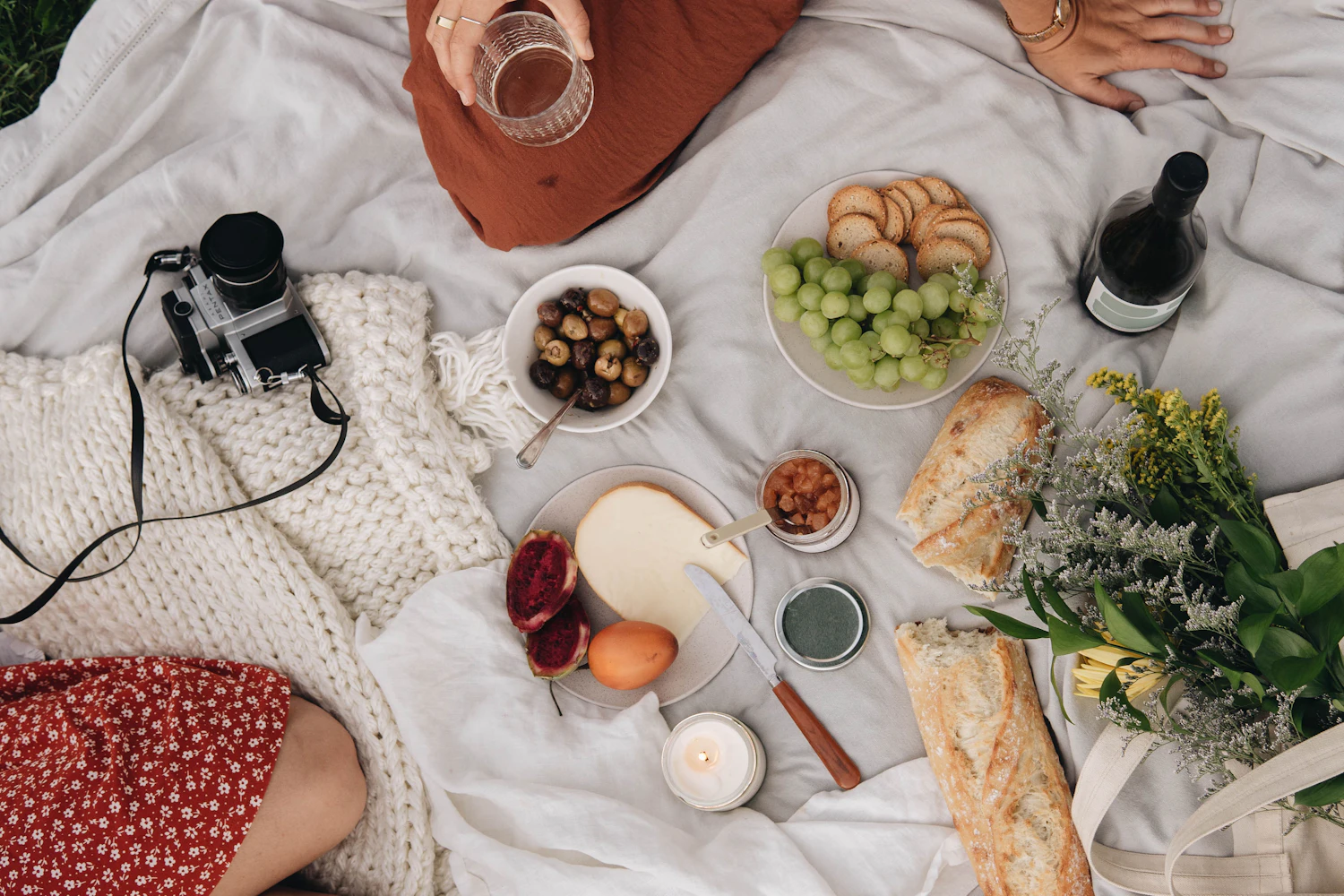 Seen from above: a white linen blanket with a baguette, grapes, a round cheese, olives and a bottle, two friends sitting at its edge