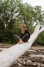 A person with long hair is sitting on a large fallen tree trunk in a natural setting. They are barefoot and dressed in casual clothing, surrounded by lush greenery and rocks, with a background of forest trees.