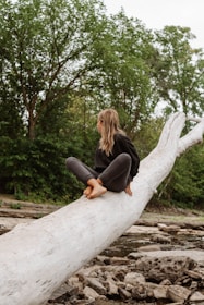 A person with long hair is sitting on a large fallen tree trunk in a natural setting. They are barefoot and dressed in casual clothing, surrounded by lush greenery and rocks, with a background of forest trees.