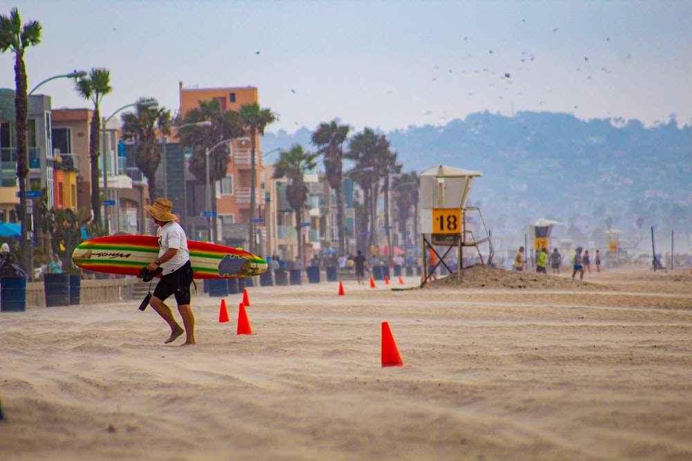 man in red shirt carrying yellow and red surfboard walking on brown sand during daytime