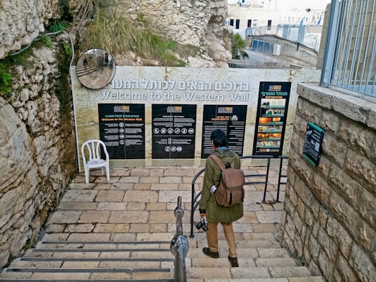 A person with a backpack stands before informational signs at the entrance to the Western Wall, Jerusalem. Stone steps and walls surround the area, with a plastic chair to the side. A convex mirror is mounted on the wall, reflecting part of the scene.