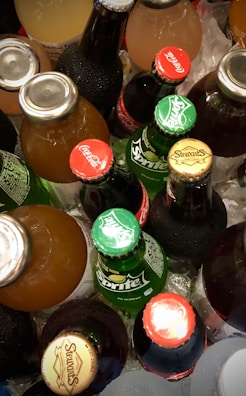 A collection of various bottled beverages placed on ice, featuring brands like Coca-Cola, Sprite, and Stewart’s. The bottles display vibrant caps with brand logos.