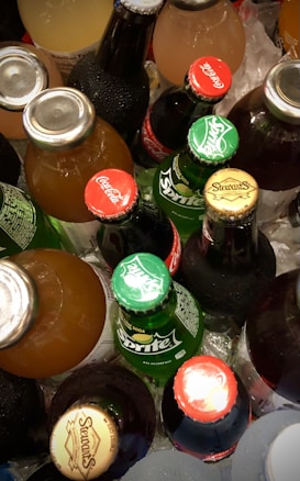 A collection of various bottled beverages placed on ice, featuring brands like Coca-Cola, Sprite, and Stewart’s. The bottles display vibrant caps with brand logos.