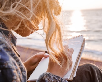 woman in blue and white floral shirt holding book near body of water during daytime