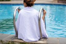 woman in white robe sitting on concrete bench near swimming pool during daytime