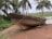 An old, weathered wooden boat is positioned on the shore, surrounded by lush greenery and palm trees. The boat is propped up with logs and appears to be undergoing repairs or maintenance. The background features a calm body of water, possibly a river or lake, under a cloudy sky.