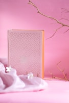 A book with a textured cover and intricate patterns stands upright against a soft pink background. Nearby, delicate white blossoms and a few branches add a natural element to the composition.