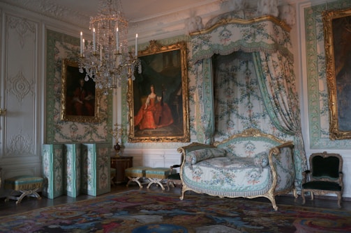 An opulent room showcasing ornate decor with intricate floral patterns. A chandelier hangs from the ceiling, illuminating the space, while a grand gilded chair and a framed portrait contribute to the luxurious atmosphere. The walls are adorned with elaborate moldings and tapestries.