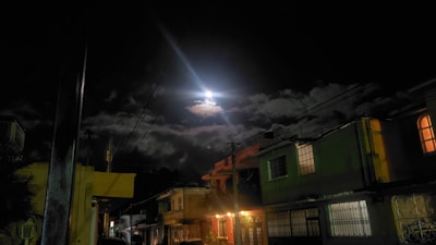 A nighttime scene featuring a street lined with houses. The bright moon is partially obscured by clouds, casting a soft glow over the area. Streetlights illuminate the buildings, highlighting their architectural details. Power lines stretch across the sky.