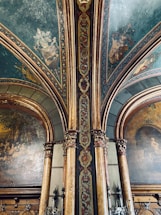 An ornate interior featuring detailed wall paintings and decorative architectural elements. The ceiling and columns are adorned with elaborate patterns and coats of arms, with a color palette of greens, golds, and deep reds. The paintings depict historical or classical scenes set within arched panels.