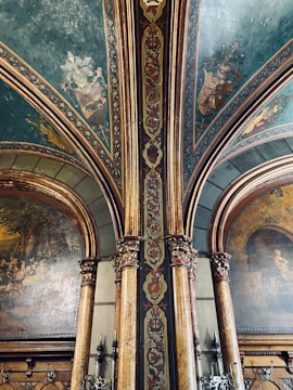 An ornate interior featuring detailed wall paintings and decorative architectural elements. The ceiling and columns are adorned with elaborate patterns and coats of arms, with a color palette of greens, golds, and deep reds. The paintings depict historical or classical scenes set within arched panels.