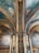 An ornate interior featuring detailed wall paintings and decorative architectural elements. The ceiling and columns are adorned with elaborate patterns and coats of arms, with a color palette of greens, golds, and deep reds. The paintings depict historical or classical scenes set within arched panels.