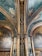 An ornate interior featuring detailed wall paintings and decorative architectural elements. The ceiling and columns are adorned with elaborate patterns and coats of arms, with a color palette of greens, golds, and deep reds. The paintings depict historical or classical scenes set within arched panels.