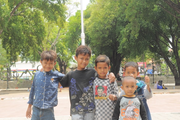 Five young children are posing together outdoors in a park-like setting with trees in the background. They appear to be standing close to each other, and some have their arms around each other. Each child wears casual clothing with various colored shirts.