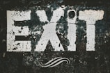 The word 'EXIT' is prominently displayed in large, bold letters that appear cracked and weathered. The paint seems to be peeling off a dark background, giving the sign an aged and rustic look. A swirling design is visible beneath the letters.