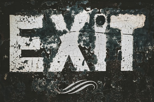The word 'EXIT' is prominently displayed in large, bold letters that appear cracked and weathered. The paint seems to be peeling off a dark background, giving the sign an aged and rustic look. A swirling design is visible beneath the letters.