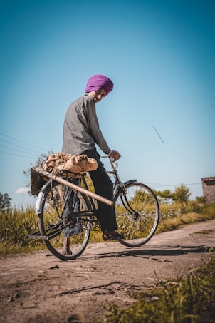 A person dressed in traditional attire and wearing a vibrant purple turban is riding a bicycle on a rural dirt path. The landscape is bright with clear blue skies and greenery lining the path. The person is carrying a bag on the back of the bicycle.