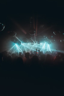 A dimly lit nightclub filled with a crowd of people dancing and enjoying themselves, illuminated by vibrant blue and green laser lights. Reflective disco balls hang from the ceiling, adding to the festive atmosphere.