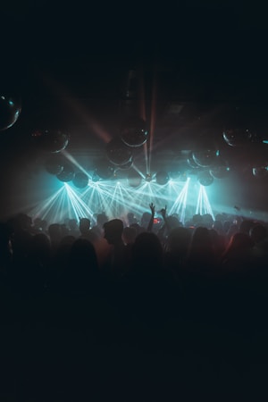 A dimly lit nightclub filled with a crowd of people dancing and enjoying themselves, illuminated by vibrant blue and green laser lights. Reflective disco balls hang from the ceiling, adding to the festive atmosphere.