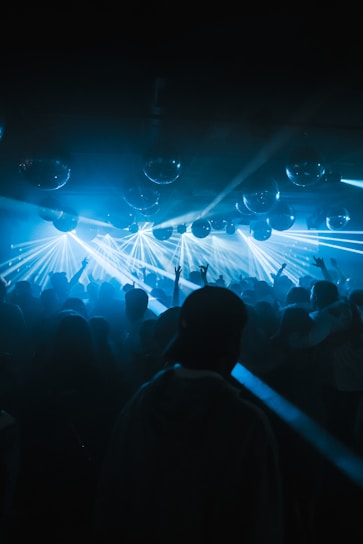 A dynamic nightclub scene filled with a dense crowd of people dancing under a ceiling adorned with disco balls. Bright blue laser lights cut across the room, illuminating the silhouettes of attendees raising their arms enthusiastically.