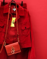 A vibrant red jacket is hanging on a red background, accompanied by a matching red handbag with a gold emblem and braided strap. The bright yellow clothing tag is prominently displayed.