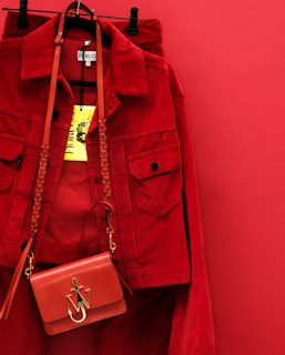 A vibrant red jacket is hanging on a red background, accompanied by a matching red handbag with a gold emblem and braided strap. The bright yellow clothing tag is prominently displayed.