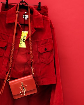 A vibrant red jacket is hanging on a red background, accompanied by a matching red handbag with a gold emblem and braided strap. The bright yellow clothing tag is prominently displayed.