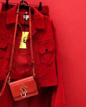 A vibrant red jacket is hanging on a red background, accompanied by a matching red handbag with a gold emblem and braided strap. The bright yellow clothing tag is prominently displayed.
