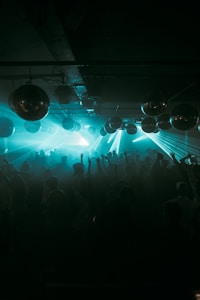 A crowded nightclub scene filled with people dancing under the glow of teal and blue lights. The atmosphere is energetic, with several disco balls hanging from the ceiling reflecting light throughout the space.