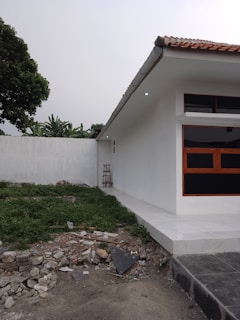 A modern, single-story white building with a red-tiled roof is adjacent to a grassy area, which contains some debris. A small ladder is positioned against the side of the building. The ground is partially covered with green vegetation and scattered construction materials near a stone wall.