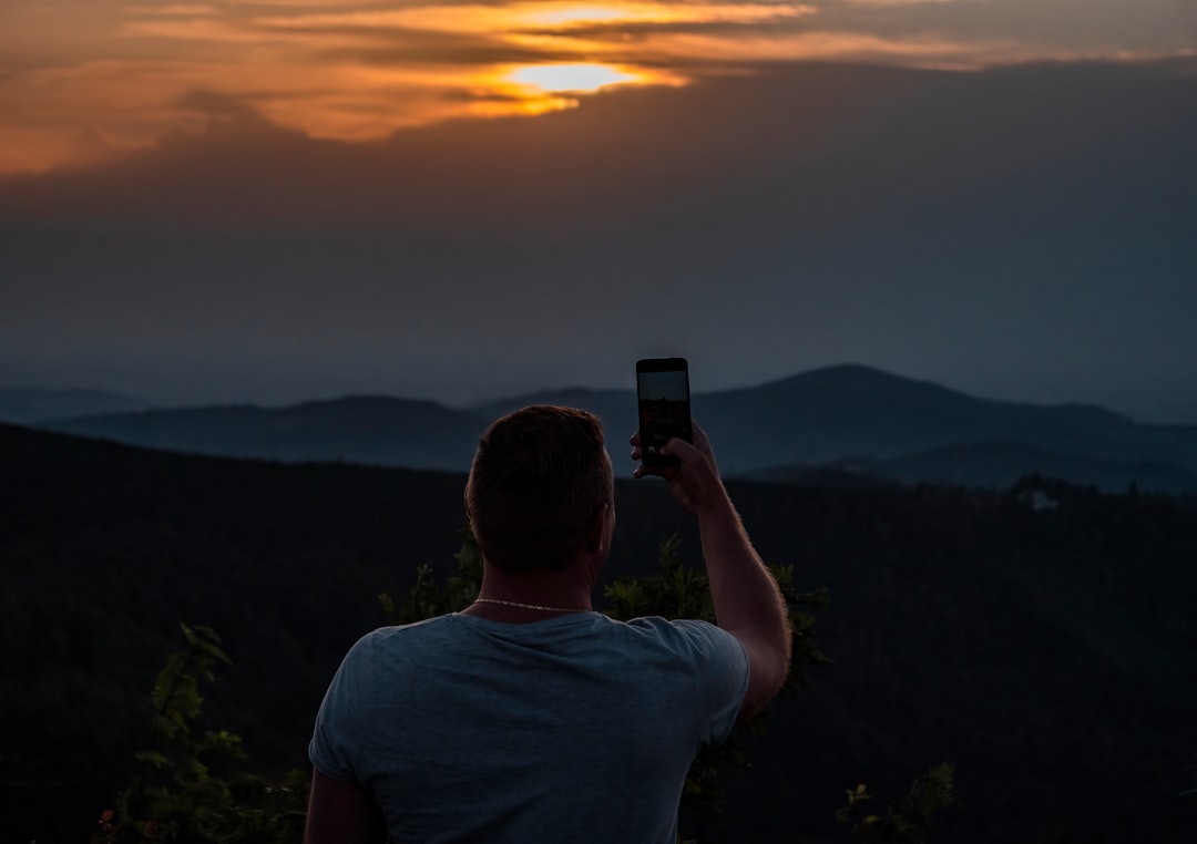man in white crew neck t-shirt holding black smartphone taking photo of mountain during sunset