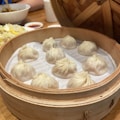 A bamboo steamer containing nine neatly arranged soup dumplings, known as xiao long bao. The dumplings have delicate pleats on their tops and rest on a paper lining. In the background, there is a serving of fried rice with diced vegetables and a bowl, along with a sauce dish.
