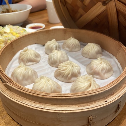 A bamboo steamer containing nine neatly arranged soup dumplings, known as xiao long bao. The dumplings have delicate pleats on their tops and rest on a paper lining. In the background, there is a serving of fried rice with diced vegetables and a bowl, along with a sauce dish.