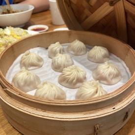 A bamboo steamer containing nine neatly arranged soup dumplings, known as xiao long bao. The dumplings have delicate pleats on their tops and rest on a paper lining. In the background, there is a serving of fried rice with diced vegetables and a bowl, along with a sauce dish.