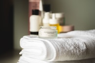 A small jar of regenerating collagen moisturizer is placed on a neatly folded, white towel. In the background, there are several blurred skincare bottles in various shapes and sizes.