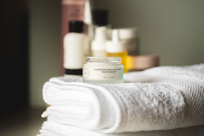 A small jar of regenerating collagen moisturizer is placed on a neatly folded, white towel. In the background, there are several blurred skincare bottles in various shapes and sizes.