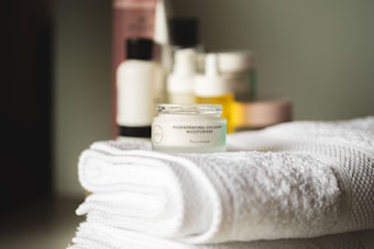 A small jar of regenerating collagen moisturizer is placed on a neatly folded, white towel. In the background, there are several blurred skincare bottles in various shapes and sizes.