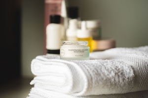 A small jar of regenerating collagen moisturizer is placed on a neatly folded, white towel. In the background, there are several blurred skincare bottles in various shapes and sizes.